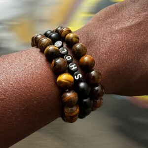 A dark-skinned wrist wearing the Ayofe 3-in-1 Customized Bracelets, featuring tiger eye beads, matte black stones, and personalized name beads spelling “SHOLA,” against a blurred background.